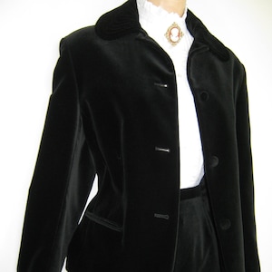 May include: A black velvet suit comprising a jacket with a notched collar and three buttons, and matching trousers. The jacket has a velvet collar and pocket trim. A white blouse and cameo brooch are visible beneath the jacket.