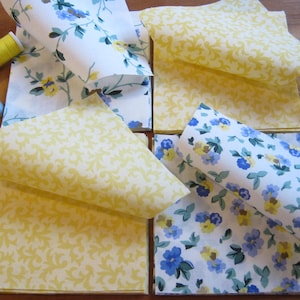 Puede incluir: Una colección de piezas de tela cuadradas con dos patrones distintos: un fondo blanco con estampados florales azules y amarillos y un fondo amarillo sólido con un diseño de enredadera blanca, junto a dos carretes de hilo.