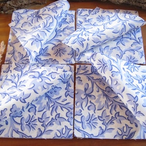 Peut inclure: Quatre dessous de verre carrés en tissu avec un fond blanc et un motif floral et végétal bleu répétitif. Chaque dessous de verre présente un coin replié, révélant le même imprimé botanique bleu et blanc sur l'envers du tissu.
