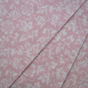 Pode incluir: Close-up de tecido rosa com um padr&atilde;o floral branco. O tecido est&aacute; dobrado, revelando os detalhes intrincados do padr&atilde;o. O design apresenta flores e folhas estilizadas, criando uma est&eacute;tica delicada e vintage.