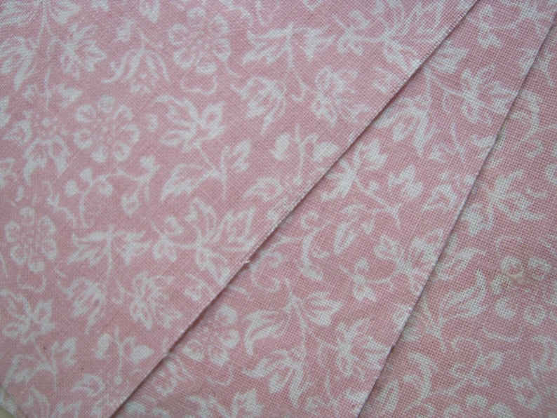 Pode incluir: Close-up de tecido rosa com um padr&atilde;o floral branco. O tecido est&aacute; dobrado, revelando os detalhes intrincados do padr&atilde;o. O design apresenta flores e folhas estilizadas, criando uma est&eacute;tica delicada e vintage.