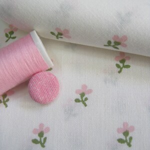 May include: A close-up of white fabric with a repeating pattern of small pink flowers and green leaves. A spool of pink thread and a pink fabric-covered button are on the fabric, suggesting sewing or crafting supplies.