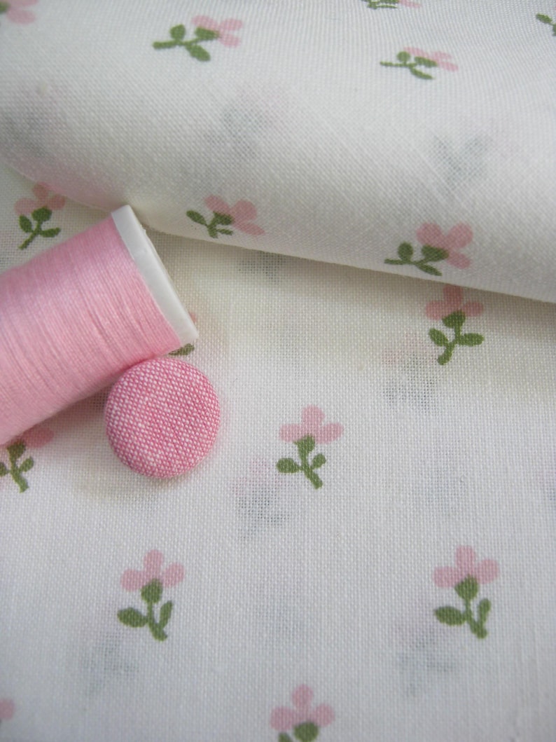 May include: A close-up of white fabric with a repeating pattern of small pink flowers and green leaves. A spool of pink thread and a pink fabric-covered button are on the fabric, suggesting sewing or crafting supplies.