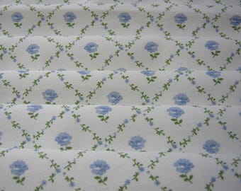 May include: A white fabric featuring a repeating pattern of light blue flowers arranged in a diamond grid. The design includes thin green vines with small leaves connecting the blue floral blossoms across the surface of the folded material.
