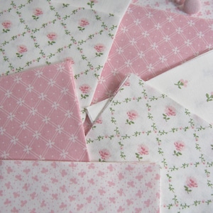 Pode incluir: Variedade de quadrados de tecido floral rosa e branco. Os tecidos apresentam v&aacute;rios padr&otilde;es, incluindo rosas, vinhas e desenhos geom&eacute;tricos. Uma bobina de linha rosa e um bot&atilde;o s&atilde;o vis&iacute;veis no fundo, sugerindo um projeto de costura ou colcha.