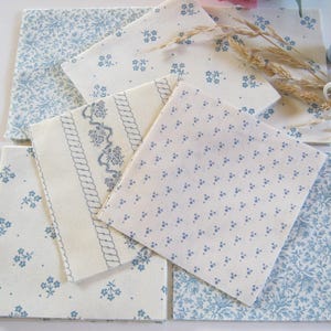 Puede incluir: Varios cuadrados de tela con diseños florales y estampados en azul claro y blanco. También se ven dos carretes de hilo azul claro, espigas de trigo secas y dos botones cubiertos de tela azul.