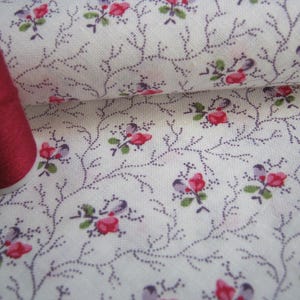 May include: A spool of dark red thread next to a white fabric with a floral pattern. The fabric features small red and purple flowers with green leaves, connected by thin, dotted purple vines.