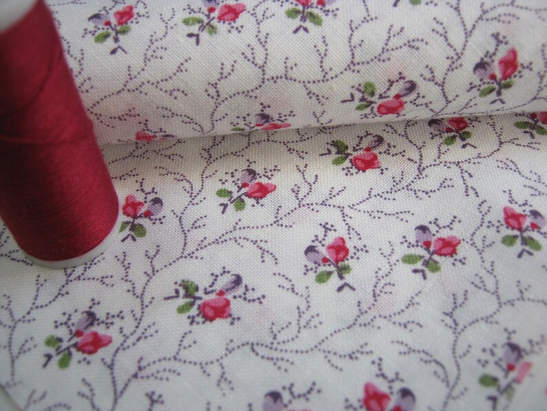 May include: A spool of dark red thread next to a white fabric with a floral pattern. The fabric features small red and purple flowers with green leaves, connected by thin, dotted purple vines.