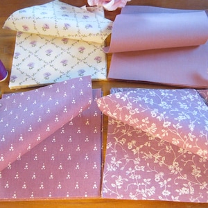 Op de afbeelding: Een verzameling van vierkante stoffen in mauve en crèmetinten. Eén lapje heeft een crèmekleurige achtergrond met een mauve bloemenpatroon, terwijl de andere mauve stof met witte bloemen- of geometrische prints tonen. Een klosje mauve garen en twee donkere knopen zijn inbegrepen.
