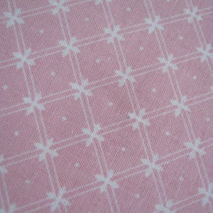Pode incluir: Close-up de um tecido rosa com um padr&atilde;o floral branco. O padr&atilde;o consiste em flores brancas em forma de cruz e pequenos pontos brancos, dispostos em um design semelhante a uma grade. O tecido tem uma apar&ecirc;ncia texturizada.