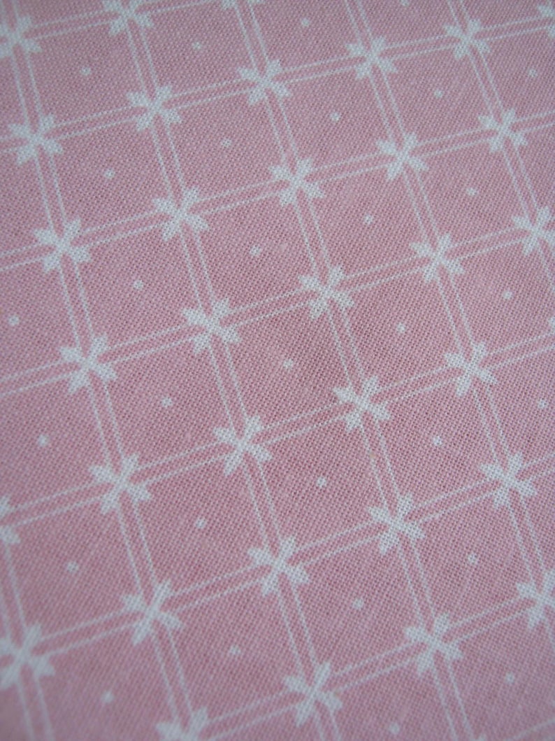 Pode incluir: Close-up de um tecido rosa com um padr&atilde;o floral branco. O padr&atilde;o consiste em flores brancas em forma de cruz e pequenos pontos brancos, dispostos em um design semelhante a uma grade. O tecido tem uma apar&ecirc;ncia texturizada.
