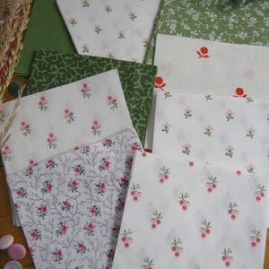 May include: Assortment of fabric squares in various floral patterns. White fabric with pink flowers, green fabric with white flowers, and green fabric with a speckled design. Also, fabric with red berries and purple floral branches.