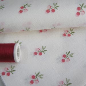 May include: White fabric with a repeating pattern of pink cherries and green leaves. A spool of red thread sits on the fabric. The fabric is folded, showing the pattern on multiple layers.