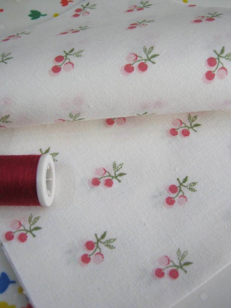 May include: White fabric with a repeating pattern of pink cherries and green leaves. A spool of red thread sits on the fabric. The fabric is folded, showing the pattern on multiple layers.