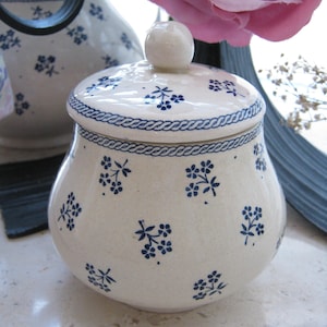 Puede incluir: Azucarero de cerámica blanca con un patrón floral azul y una tapa. El tazón tiene una forma redonda y una pequeña perilla en la tapa.