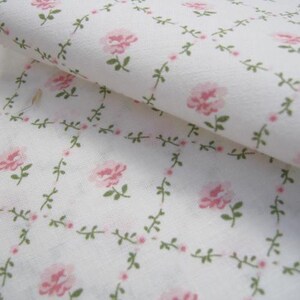 Pode incluir: Close-up de tecido branco com um padr&atilde;o repetido de rosas cor-de-rosa e folhas verdes. O desenho floral &eacute; disposto em um padr&atilde;o de grade com pequenos pontos cor-de-rosa. O tecido est&aacute; dobrado, mostrando o padr&atilde;o nas camadas superior e inferior.