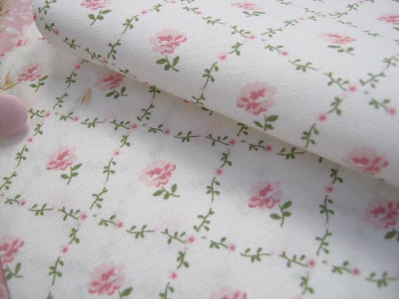Pode incluir: Close-up de tecido branco com um padr&atilde;o repetido de rosas cor-de-rosa e folhas verdes. O desenho floral &eacute; disposto em um padr&atilde;o de grade com pequenos pontos cor-de-rosa. O tecido est&aacute; dobrado, mostrando o padr&atilde;o nas camadas superior e inferior.