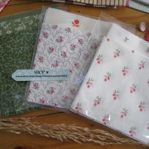 May include: Several fabric bundles in clear packaging, featuring floral and botanical patterns in green, white, and pink. A light blue tag with the word "SHOP" and a website address is visible. The fabrics are arranged on a wooden surface.