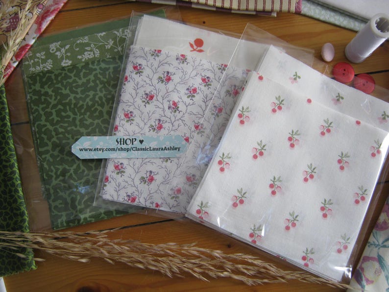 May include: Several fabric bundles in clear packaging, featuring floral and botanical patterns in green, white, and pink. A light blue tag with the word "SHOP" and a website address is visible. The fabrics are arranged on a wooden surface.