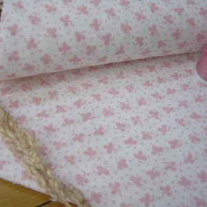 Pode incluir: Close-up de tecido branco com padr&atilde;o de trevos rosa, parcialmente enrolado para revelar o verso. Uma bobina de linha rosa est&aacute; ao lado. Dois bot&otilde;es revestidos de tecido e uma espiga de trigo seca tamb&eacute;m s&atilde;o vis&iacute;veis.