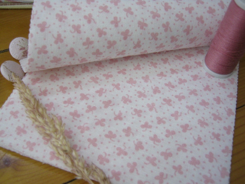 Pode incluir: Close-up de tecido branco com padr&atilde;o de trevos rosa, parcialmente enrolado para revelar o verso. Uma bobina de linha rosa est&aacute; ao lado. Dois bot&otilde;es revestidos de tecido e uma espiga de trigo seca tamb&eacute;m s&atilde;o vis&iacute;veis.