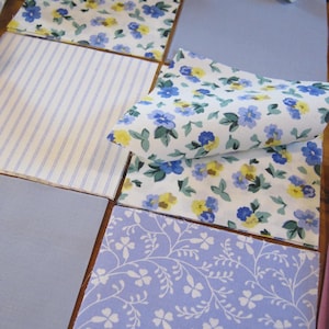 Puede incluir: Una colección de cuadrados de tela con varios patrones, incluyendo un fondo blanco con estampados florales azules y amarillos, un diseño de rayas finas azul claro y blanco, una tela azul claro sólida y una tela morada clara con patrones de enredaderas blancas.