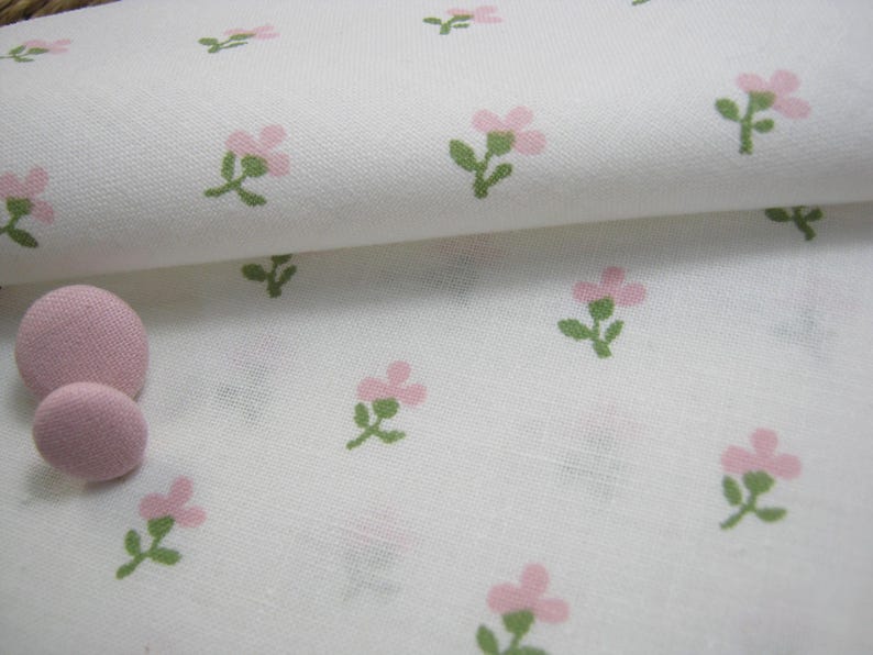 Pode incluir: Tecido branco com um padr&atilde;o repetido de pequenas flores rosa e caules verdes. Dois bot&otilde;es rosa revestidos de tecido s&atilde;o vis&iacute;veis. O tecido tem uma apar&ecirc;ncia ligeiramente texturizada, sugerindo uma fibra natural como algod&atilde;o ou linho.