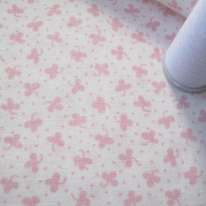 Pode incluir: Tecido branco com um padr&atilde;o repetitivo de borboletas rosa e pequenos pontos. Um rolo de linha branca &eacute; vis&iacute;vel no canto. O tecido tem uma textura, possivelmente algod&atilde;o ou linho.