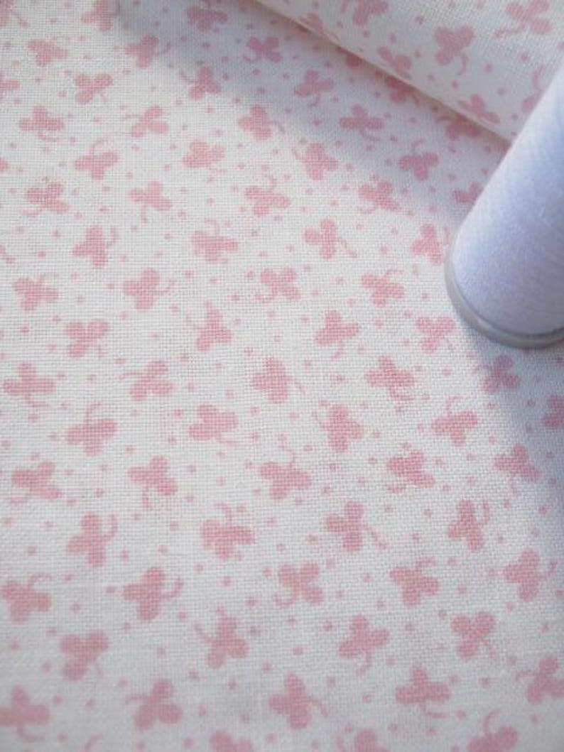 Pode incluir: Tecido branco com um padr&atilde;o repetitivo de borboletas rosa e pequenos pontos. Um rolo de linha branca &eacute; vis&iacute;vel no canto. O tecido tem uma textura, possivelmente algod&atilde;o ou linho.