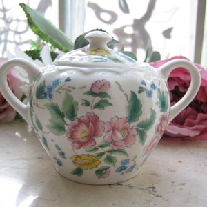 Könnte beinhalten: Weiße Porzellan-Zuckerdose mit floralem Design mit rosa, gelben, blauen und grünen Blumen. Der Deckel hat einen Knopf mit einer gelben Blume.