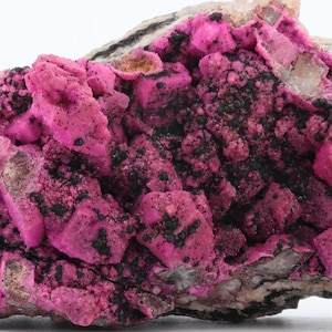 May include: A natural mineral specimen with a striking colour contrast. The rock has a grey and black base, with a prominent cluster of bright pink, crystalline formations. The overall texture is rough and uneven, showcasing natural geological beauty.