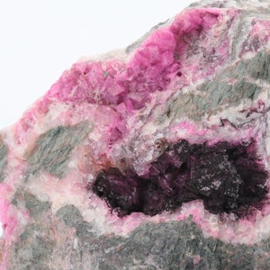 May include: A natural rock formation with a striking contrast of colours. The rock features bright pink crystalline formations and a dark, cave-like interior. The exterior shows grey and pink hues, set against a white backdrop.