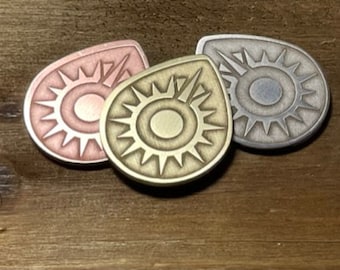Puede incluir: Tres fichas de metal con un diseño de sol. Las fichas son de diferentes colores: oro rosa, plata y latón.