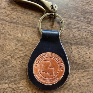 May include: A black leather keychain with a brown leather circle featuring the text "The Boot Leather Co." and an outline of the state of Louisiana. The keychain has a brass hook.