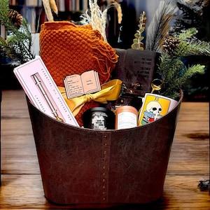May include: A brown leather basket filled with a variety of gifts, including a  candle, a book, a diffuser pen, and a tea bag. The basket is sitting on a wooden table.