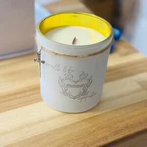 May include: A white candle in a glass jar with a gold rim. The jar has a decorative cross charm and the words "smells like" and "Fireheart Halo" printed on it. The candle sits on a wooden surface.