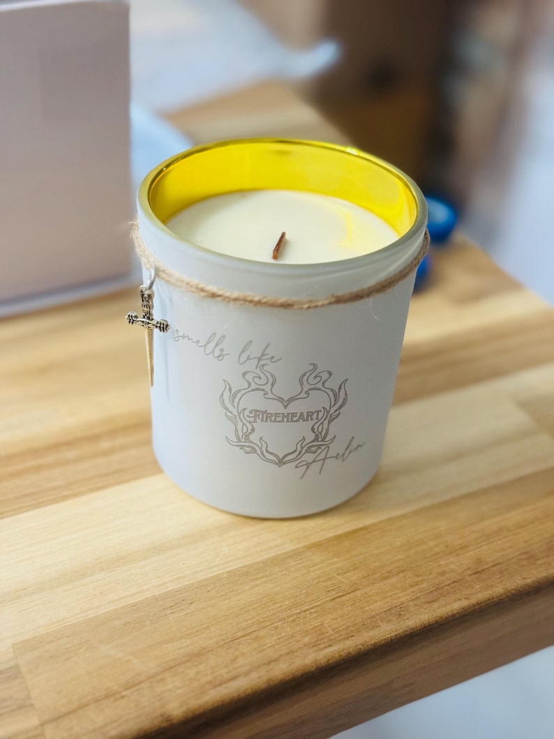 May include: A white candle in a glass jar with a gold rim. The jar has a decorative cross charm and the words "smells like" and "Fireheart Halo" printed on it. The candle sits on a wooden surface.