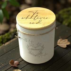 May include: A white candle jar with a wooden lid, featuring the text "To whatever end." The jar is adorned with a decorative rope and a silver charm. The jar also has the text "smells like Fireheart" and a heart design.