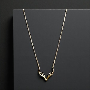 May include: A gold-colored necklace featuring a pendant shaped like deer antlers. The delicate chain and antler pendant are displayed against a dark gray background. The necklace is a minimalist piece of jewelry.