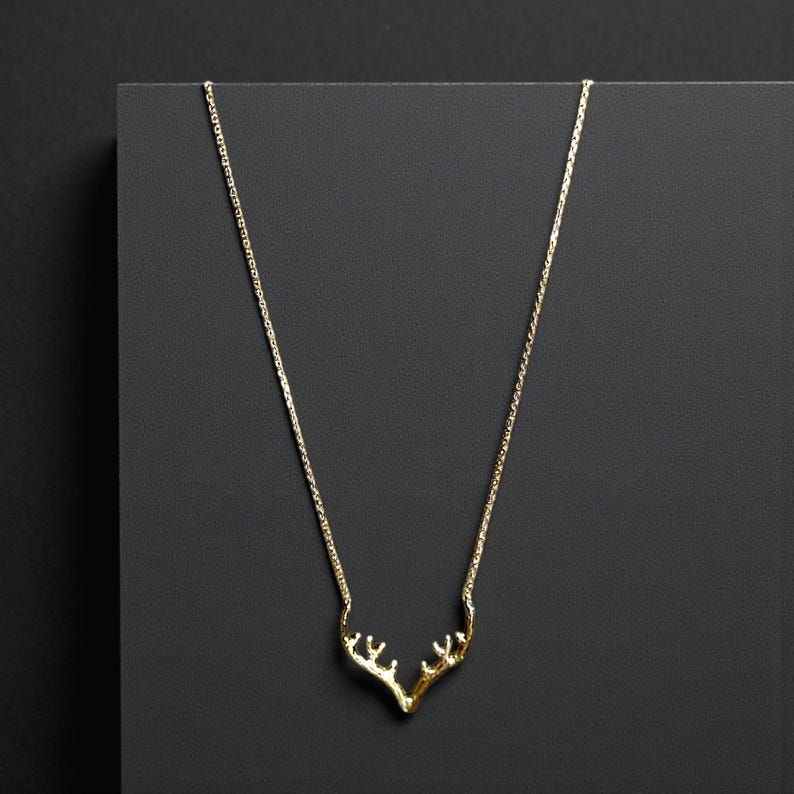 May include: A gold-colored necklace featuring a pendant shaped like deer antlers. The delicate chain and antler pendant are displayed against a dark gray background. The necklace is a minimalist piece of jewelry.