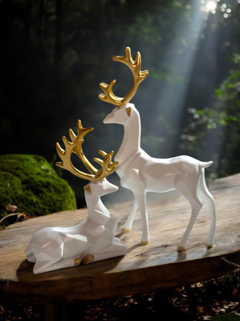 May include: Two white deer figurines with gold antlers are displayed on a wooden surface. One deer is standing, and the other is lying down. The background is a blurred forest scene with sunlight.