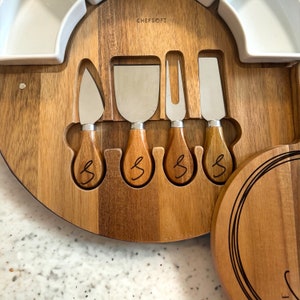 K&ouml;nnte beinhalten: Ein K&auml;sebrett-Set aus Holz mit vier K&auml;semessern aus Edelstahl. Die Messer haben Holzgriffe mit dem eingravierten Buchstaben "S". Das Brett hat einen wei&szlig;en Keramikbereich und die Marke "CHEFSOFI" ist sichtbar.
