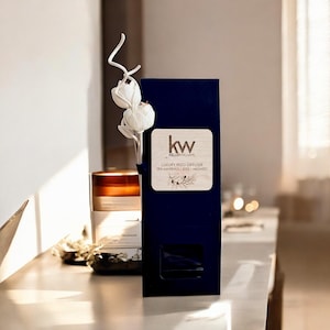 May include: A navy blue box with a wooden label featuring the "kw" logo and text for a luxury reed diffuser. A white floral arrangement and a candle are visible. The diffuser box has a small window.