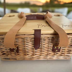 May include: A wicker picnic basket with a wooden lid. The lid has two leather straps and a brown leather handle. The lid is engraved with the words "Bran" and "Travel" in cursive.