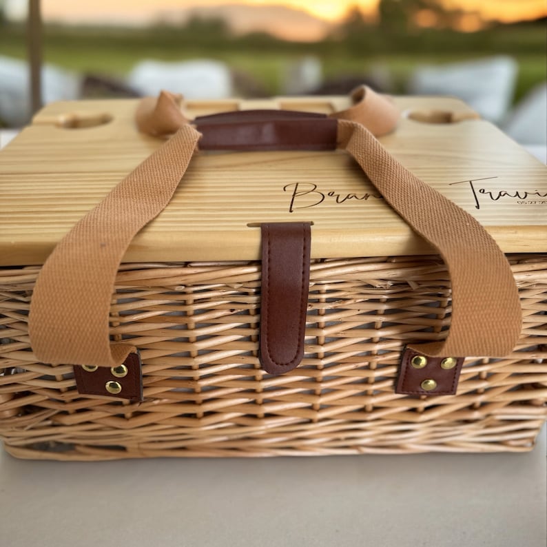 May include: A wicker picnic basket with a wooden lid. The lid has two leather straps and a brown leather handle. The lid is engraved with the words "Bran" and "Travel" in cursive.