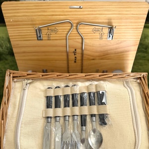 May include: A wicker picnic basket with a wooden lid and a set of silverware inside. The silverware is wrapped in black and white plastic and includes a knife, fork, spoon, and a butter knife. The lid has a metal hook and the text "Hold Down" printed on it.