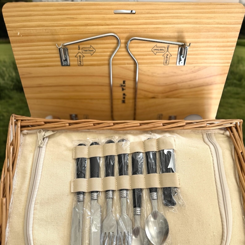 May include: A wicker picnic basket with a wooden lid and a set of silverware inside. The silverware is wrapped in black and white plastic and includes a knife, fork, spoon, and a butter knife. The lid has a metal hook and the text "Hold Down" printed on it.