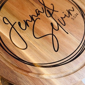 May include: A round wooden serving board with a black engraved circle containing the text "Jenna & Kevin 5.5.24". The board is part of a cheese board set with a knife and a fork.