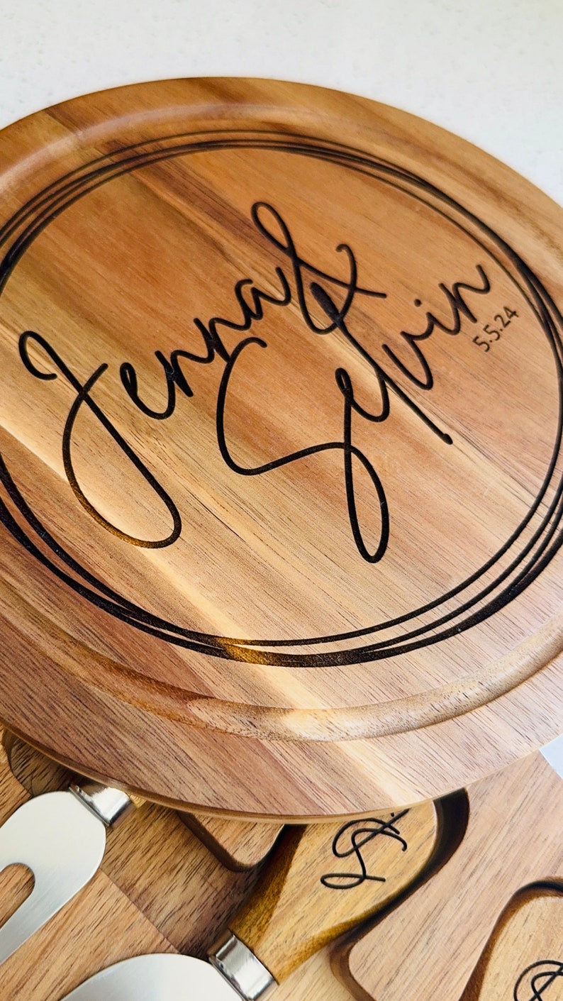 May include: A round wooden serving board with a black engraved circle containing the text "Jenna & Kevin 5.5.24". The board is part of a cheese board set with a knife and a fork.