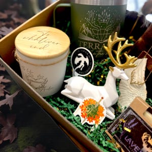 May include: A gift box filled with various items, including a white candle with a wooden lid, a green mug with a deer design, a white deer figurine with gold antlers, and stickers. The box is lined with green paper.